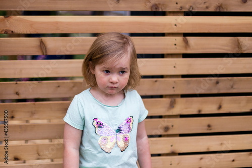 Horizontal medium view of cute blond toddler girl standing in front of wooden fence with dejected expression 