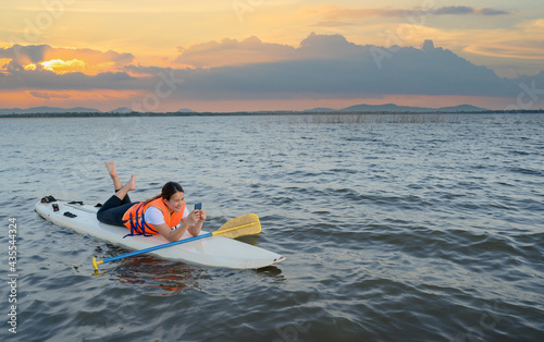 Asian athletic woman with mobile phone on stand paddle board in lake. Solo outdoor SUP activity on summer holiday.