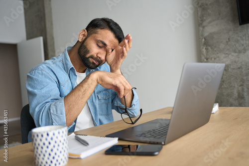 Young indian eastern tired exhausted business man rubbing eyes sitting in modern home office with laptop on desk. Overworked burnout academic Hispanic student with glasses in hand feeling eyestrain.