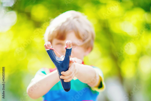 Funny little kid boy shooting wooden slingshot against green tree background. Child having fun in summer