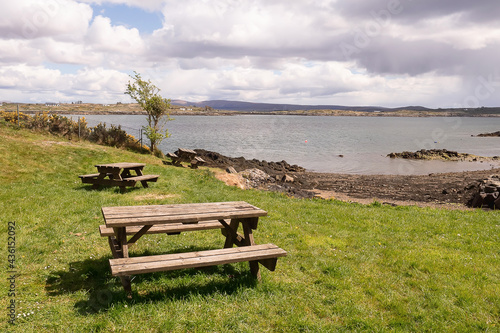 Picnic area by the ocean with beautiful view. Wooden benches and tables on the grass. Warm sunny day. Roundstone town, county Galway, Ireland. Nobody.