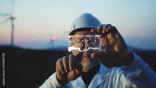 Adult man scientist using high tech smart phone to diagnose online the workflow on modern windmill farm. Beautiful sunset sky on background. The newest technology trend of communication.