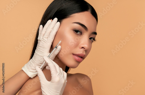 Young Asian woman with clean radiant skin gets botox injections for contour tightening, lip augmentation on a beige background. Spa care, facial skin care, beauty cosmetology.