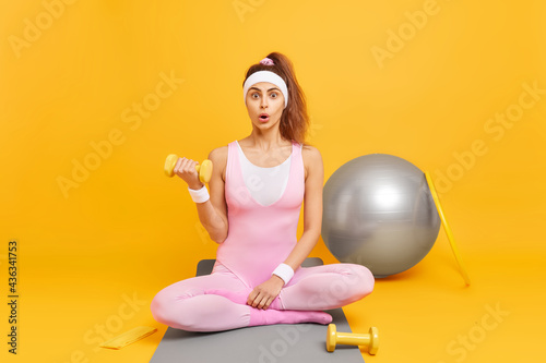 Full length shot of surprised woman has fitness practice lifts dumbbell poses crossed legs on karemat does exercises in sports club isolated over yellow background. People wellness and sport