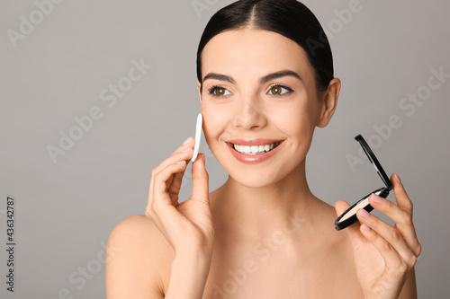 Beautiful young woman applying face powder with puff applicator on grey background