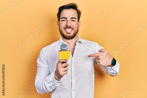 Handsome caucasian man with beard holding reporter microphone pointing finger to one self smiling happy and proud