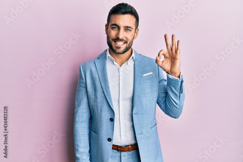 Young hispanic businessman wearing business jacket smiling positive doing ok sign with hand and fingers. successful expression.