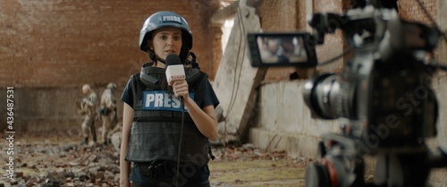 Behind the scenes of female war journalist correspondent wearing bulletproof vest and helmet reporting live near destroyed building, military personnel in the background