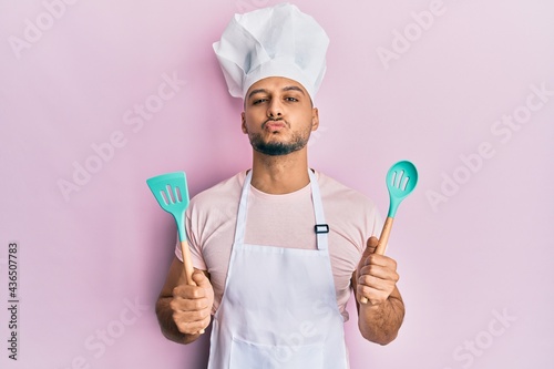 Young arab man wearing professional cook apron and hat holding spoon looking at the camera blowing a kiss being lovely and sexy. love expression.