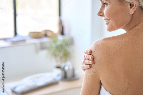 Back view of nude beautiful middle aged 50s woman applying exfoliating peeling sugar scrub after shower. Advertising of bodycare professional spa therapy at home using antiage bodycare products.