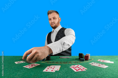 Male croupier at the casino at the table isolated on blue background. Casino concept, gambling, poker, chips on the green casino table.