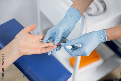 Close up of nurse lab technician in blue sterile gloves, using painless scarifier to prick the finger of unrecognizable patient, sitting on chair. Blood analysis and group determination