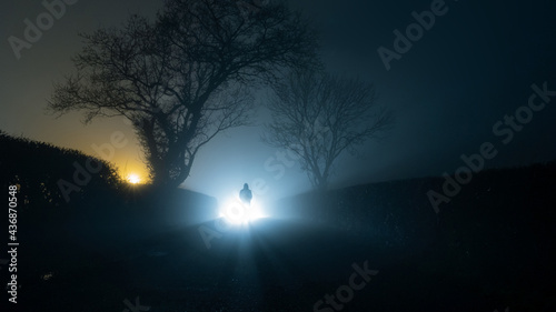 A scary, mysterious hooded figure, standing in front of a bright light on a country lane, on a spooky foggy winters night.