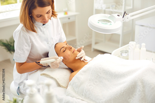 Microdermabrasion skincare in beauty salon. Smiling woman dermatologist making skincare procedure of ultrasound Microdermabrasion with special metal machine for relaxing young woman