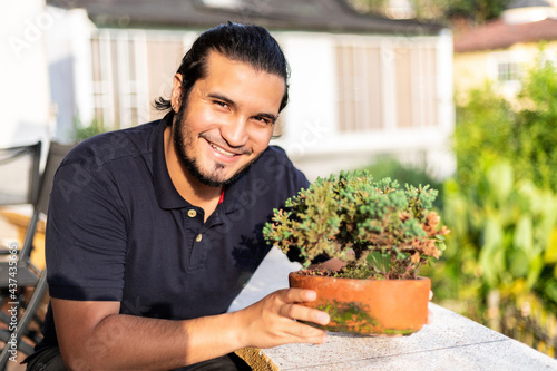 Young latino man taking care of a bonsai in a clay pot.