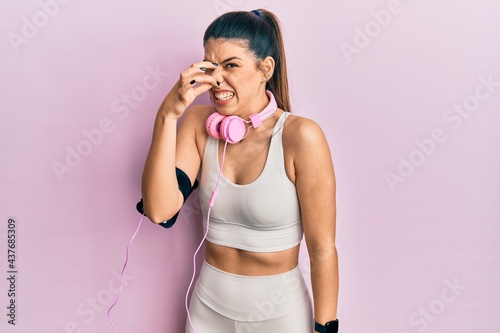 Young hispanic woman wearing gym clothes and using headphones smelling something stinky and disgusting, intolerable smell, holding breath with fingers on nose. bad smell