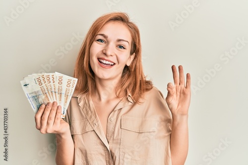 Young caucasian woman holding 100 danish krone banknotes doing ok sign with fingers, smiling friendly gesturing excellent symbol