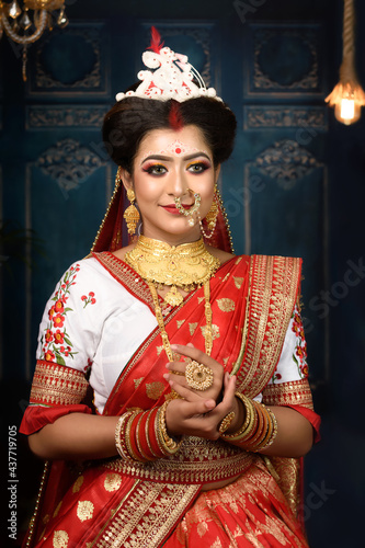 Stunning Indian bride dressed in Hindu red traditional wedding clothes sari embroidered with gold jewellery and a veil smiles tender in studio lighting indoor. Ethnic Bengali wedding.