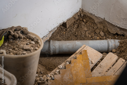 Leaking plastic sewage pipe in an old apartment, visible open hole with removed parquet, sand and buckets around, exposed drain pipe in the ground.