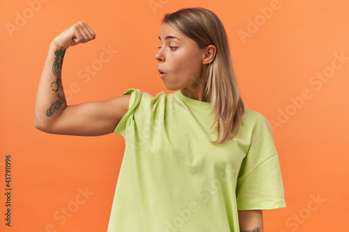 Surprised beautiful young woman in yellow tshirt showing biceps muscles and looking on it isolated over orange background