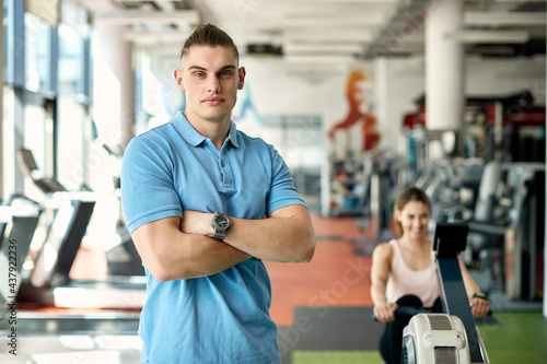 Portrait of confident personal trainer with woman exercising in the background at the gym.