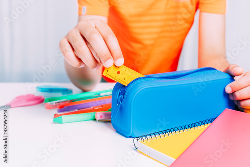 Boy keeping inside his blue pencil case all a small ruler and pencils, pencils, scissors, etc. Colorful notebooks on white table. Back to school concept