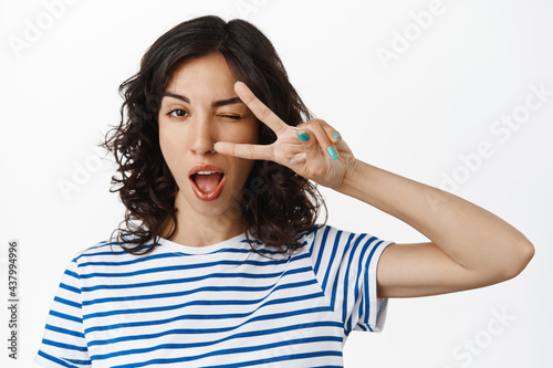 Close up portrait of positive brunette girl show peace, kawaii v-sign and tongue, winking happy at camera, standing relaxed against white background