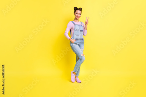 Photo of charming young girl posing show v-sign shiny toothy smile wear denim overall shoes isolated yellow background