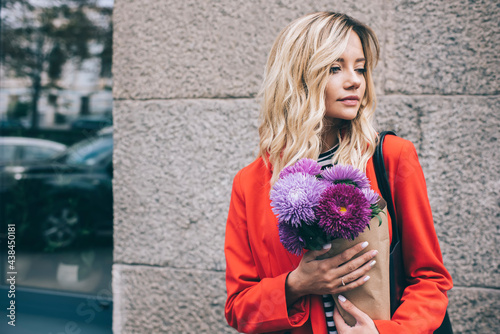 Contemplative hipster girl holding floristic present delivered in city thinking about surprise outdoors, pensive female touristic fashionista with violet flowers feeling pondering