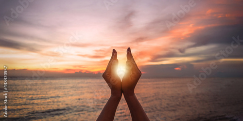June summer solstice sun concept with silhouette of happy young woman's hands relaxing, meditating and holding sunset against warm golden hour sky on the beach with natural ocean or sea background