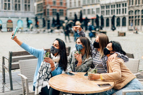Multiracial female group friends around a table in cafe terrace with drink. Centennial friends taking selfie smiling behind face masks - New normal concept