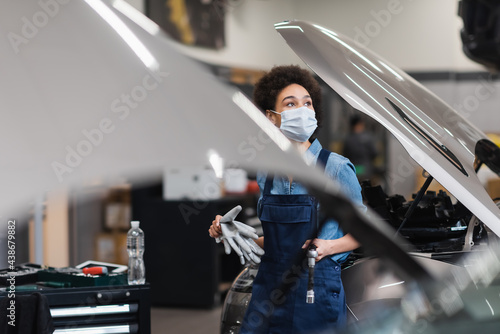 young african american mechanic in protective mask holding equipment near cars with open hoods in garage