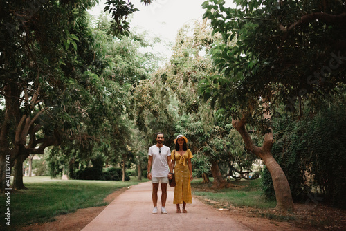 A brunette girl in a yellow dress and her boyfriend are staying on a sand path between trees in the Valencian park. A couple of tourists on a date in summer in the evening.