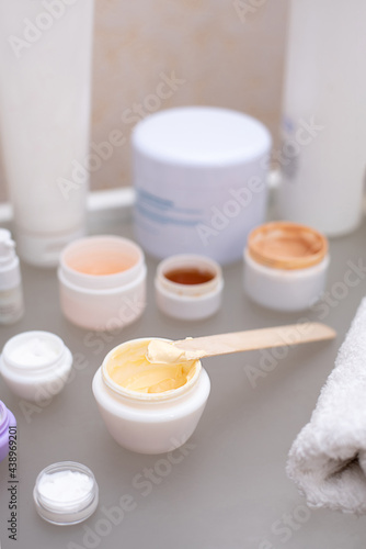 Wooden spatula with yellow cream on a jar on a glass table with bottles of cream, skin care concept.