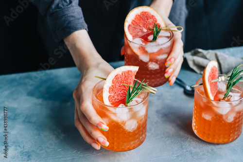 Female bartender hand holding grapefruit cocktail lemonade glasses with ice and rosemary.