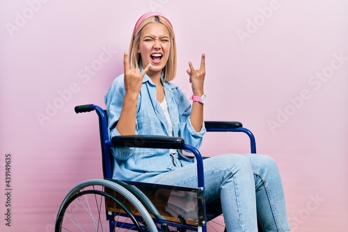 Beautiful blonde woman sitting on wheelchair shouting with crazy expression doing rock symbol with hands up. music star. heavy music concept.