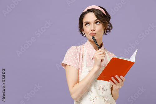 Young thoughtful housewife housekeeper chef cook baker woman in pink apron write down recipe in cook book making notes in notebook isolated on pastel violet background. Cooking food process concept