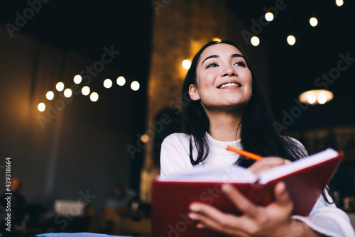 Happy Caucasian poet satisfied with creation process sitting at table with education textbook and smiling, cheerful female with brunette hair enjoying leisure time for planning week schedule