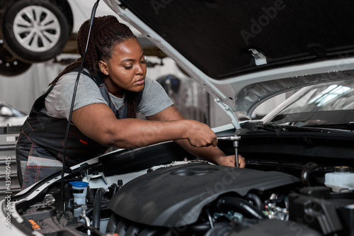 Black female mechanic working under the hood at repair garage. Portrait of confident focused mechanic woman working ona car in an auto repair shop. Female mechanic working on car. Side view