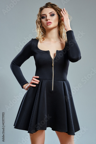 Elegant blonde woman with curly hair, bright make-up, in black stylish dress is posing against gray studio background. Fashion and beauty. Close-up.