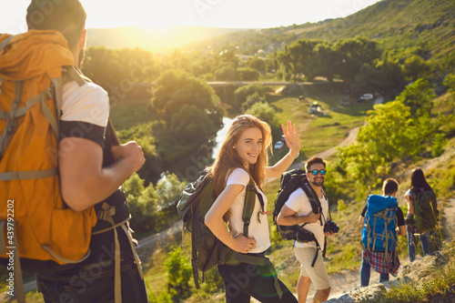 Group of happy backpackers trekking on sunny day. Young tourists traveling and enjoying active summer vacation. Smiling woman looking at camera and waving hand walking down hiking trail with friends