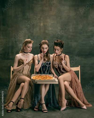 Pretty women in vintage 20s dresses. Young girls in art action sitting together and posing on wall background.