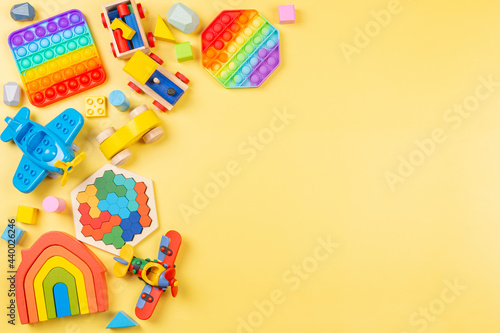 Baby kids toys background with wooden rainbow color house, train, car, plane, pop it fidget toys and colorful blocks on yellow background. Top view, flat lay