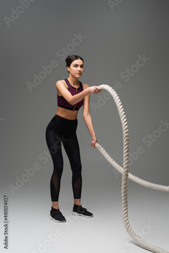 full length view of sportive woman exercising with battle ropes on grey background.