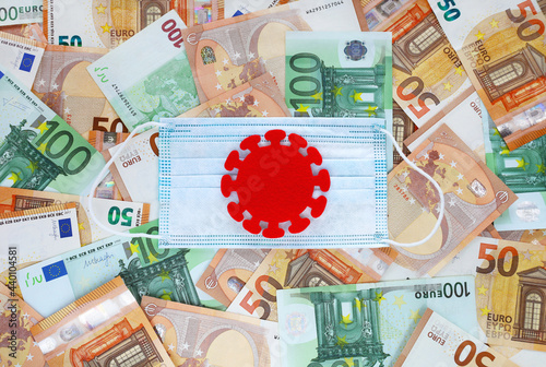 Red felt covid virus centered on blue medical mask on 50 and 100 euro banknotes background. Coronavirus pandemic in Europe. Financial crisis, bank, money, economy, business concept. Place for text.