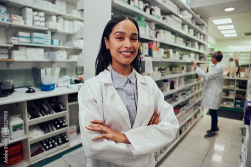 Portrait of young african american pharmacist standing between aisle in chemist with colleague working in background