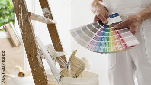 hands of house painter man decorator choose the color using the sample swatch, work of the house to renovate, a wooden ladder with paint brushes and a bucket as a background, close up