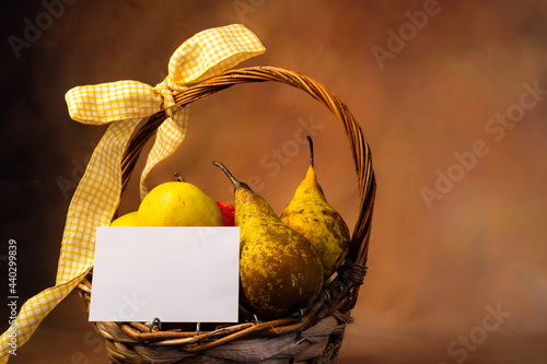 Scena con un cesto di frutta pieno di mele e pere con un biglietto di auguri bianco e legato con un fiocco giallo. Alimentazione. Salute. Invito.