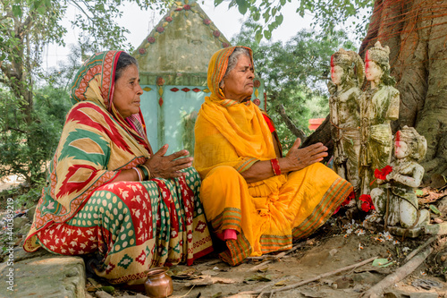 Two mature ladies from rural India background praying outdoors with folded hands and closed eyes wearing ethnic saree. They are worshipping gods of Hindu mythology viz idols of Ram, Laxman and Hanuman