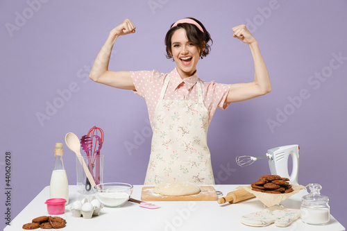 Young housewife housekeeper chef cook baker woman in pink apron work at table kitchenware show hand biceps muscles demonstrate power isolated on pastel violet background Process cooking food concept